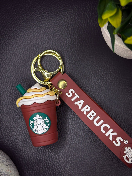 A green Starbucks frappuccino-shaped keychain with a whipped cream top and caramel drizzle, attached to a green rubber strap with the Starbucks logo, finished with a golden keyring.