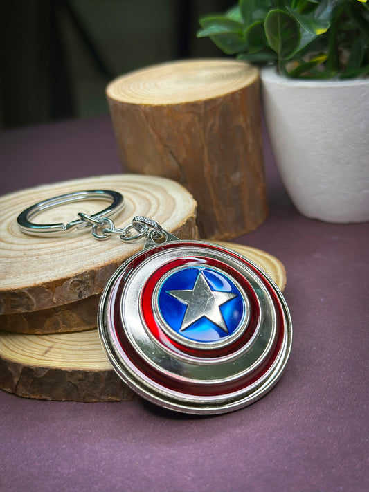 A Captain America shield-themed keychain with a metallic red, silver, and blue spinner design placed on wooden slices with a plant in the background.
