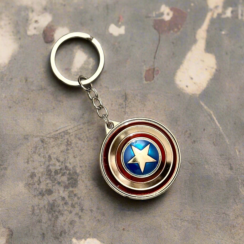 A Captain America shield-themed keychain with a metallic red, silver, and blue spinner design placed on wooden slices with a plant in the background.