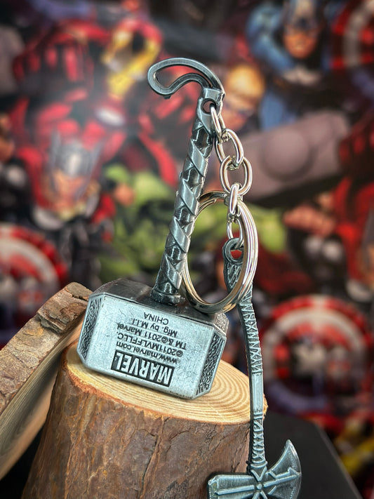 Two metallic Marvel keychains: Thor’s Mjolnir hammer and Stormbreaker axe, placed on a black surface with engraved detailing visible.