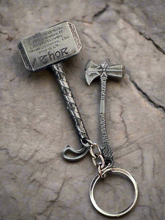 Two metallic Marvel keychains: Thor’s Mjolnir hammer and Stormbreaker axe, placed on a black surface with engraved detailing visible.