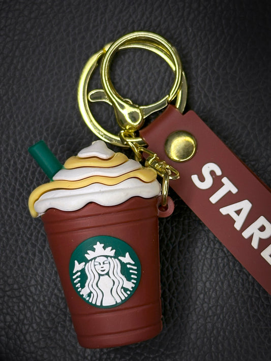 A green Starbucks frappuccino-shaped keychain with a whipped cream top and caramel drizzle, attached to a green rubber strap with the Starbucks logo, finished with a golden keyring.