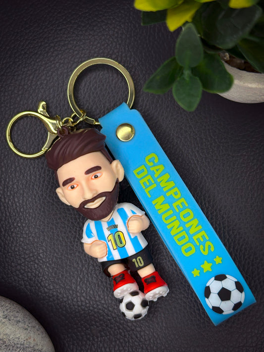 A cartoon keychain of Messi in the Argentina jersey with a football at his feet, attached to a light blue strap that reads “Campeones Del Mundo” with stars and soccer ball icons.