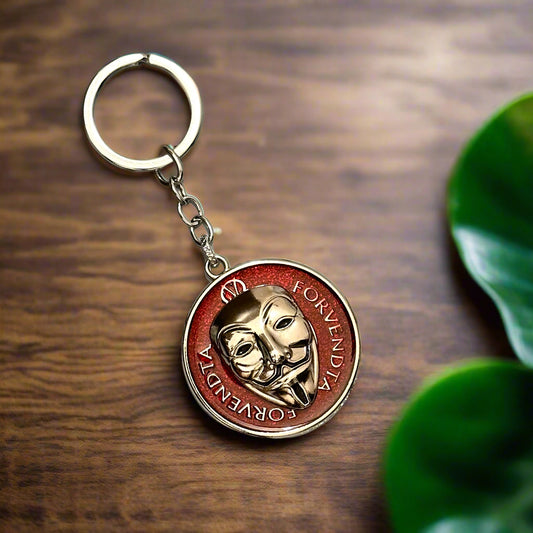 A circular keychain featuring the golden Guy Fawkes mask from V for Vendetta, encased in a red background with the text “FORVENDTA,” placed on a white marble hexagon-tiled background.