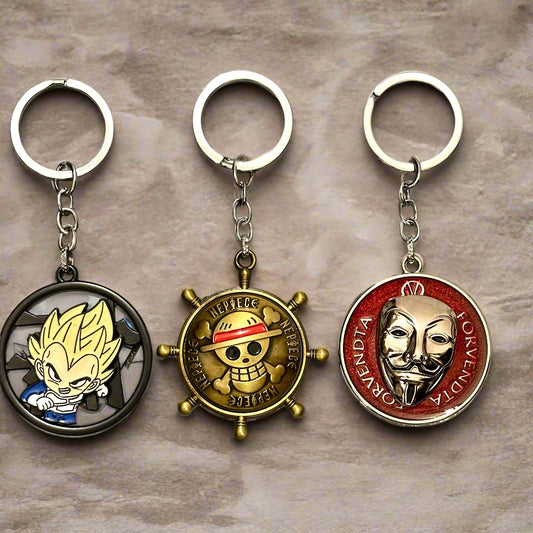 A trio of metallic spinner keychains featuring: One Piece ship wheel with Luffy’s emblem, a red V for Vendetta mask spinner, and a Dragon Ball Z Super Saiyan Goku surrounded by flames—all arranged on a white marble tile background.
