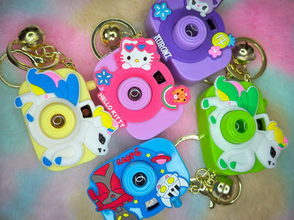 Five colorful cartoon camera keychains with characters like Hello Kitty, Kuromi, unicorns, and robots, laid out on a pastel fluffy surface. Each has a gold keyring and jingle bell.