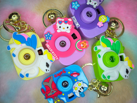 Five colorful cartoon camera keychains with characters like Hello Kitty, Kuromi, unicorns, and robots, laid out on a pastel fluffy surface. Each has a gold keyring and jingle bell.